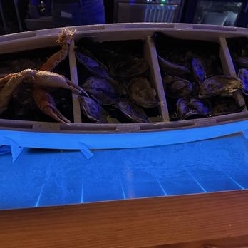 Fin N Tonic - A boat-shaped seafood display on a table contains multiple oysters arranged in sections, with a crab placed on top of the oysters. The display is illuminated with blue lighting, and the setting appears to be a restaurant or bar environment, likely specializing in fresh seafood and oysters. - Seafood Near Me - Denver, Colorado
