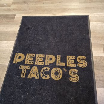 Peeples Taco’s - A black rectangular mat with yellow text that says 'PEOPLE'S TACOS' is placed on a wooden floor, with the top of a person's shoes visible at the top edge. - Fast Food Near Me - Hazel Crest, Illinois
