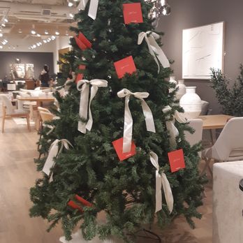 Structube - Inviting for the festive season 2025/26 - A Christmas tree decorated with white ribbons and red cards is positioned in a furniture store showroom. The store features modern furniture, artwork, and decorative items, with warm lighting and a wooden floor. The tree is placed on a white tree skirt, and the background includes various furniture pieces and wall art. - Furniture Stores Near Me - Edmonton, Alberta