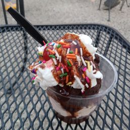 Chocolate Peanut Butter - A dessert sundae with whipped cream, colorful sprinkles, chocolate syrup, and a cherry on top, served in a clear cup on a metal outdoor table.