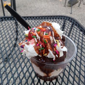 Mrs. Sprinkles Ice Cream - Vegan sundae (chocolate peanut butter and cherry chocolate chip ice cream) with vegan whipped cream and vegan choc fudge, sprinkles+cherry - A dessert sundae with whipped cream, colorful sprinkles, chocolate syrup, and a cherry on top, served in a clear cup on a metal outdoor table. - Ice Cream & Frozen Yogurt Near Me - Albuquerque, New Mexico