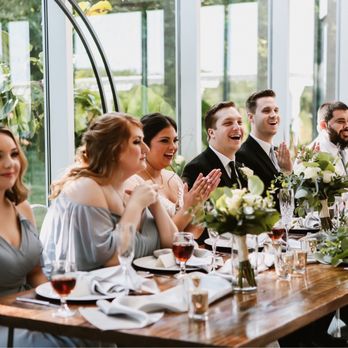 Hired Hostess Event - Six individuals are seated at a wooden table decorated with white and green floral arrangements, enjoying a celebration in a bright, glass-enclosed venue. They are dressed in formal attire, with women in dresses and men in suits, and are smiling and clapping, indicating a joyful occasion. The setting features large glass windows allowing natural light to illuminate the scene, with additional greenery visible outside. - Wedding Planning Near Me