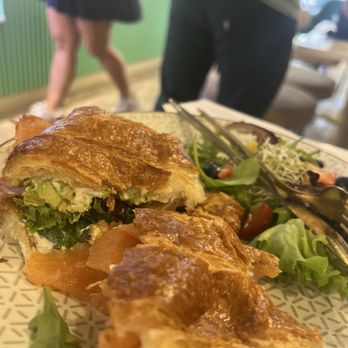 Vanilla Cafe - A close-up of a croissant sandwich containing avocado, lettuce, and smoked salmon on a patterned plate. The plate also has a side salad with mixed greens, cherry tomatoes, blueberries, and sprouts. In the background, a person in a gray shirt and dark pants is partially visible, with another person in a skirt walking nearby in a restaurant or cafe setting. - Cafes Near Me - Miami, Florida