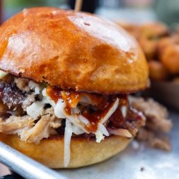 Classic BBQ Sandwich - A close-up image of a barbecue sandwich featuring shredded meat, coleslaw, and barbecue sauce inside a toasted bun. The sandwich is placed on a tray, with a bowl of fried okra visible in the background. The setting suggests a casual dining environment, likely at a barbecue restaurant.