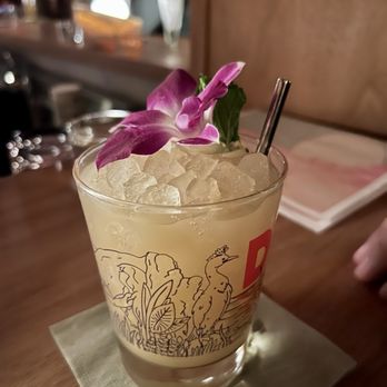 Daydream - Mai Thai - A cocktail served in a clear glass featuring a black and red illustration of a bird and plants. The drink is topped with crushed ice, a purple flower, and a green leaf, with a metal straw inserted. The glass is placed on a napkin on a wooden table, with a blurred background of a bar or restaurant setting. - Cocktail Bars Near Me - Albuquerque, New Mexico