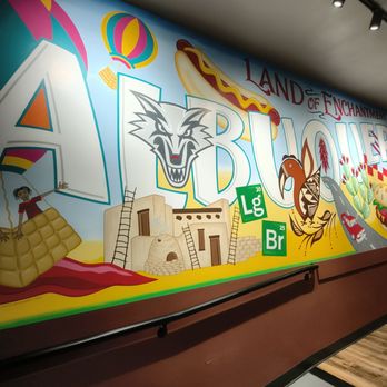 Laguna Burger - The mural displays a colorful scene with various elements including a hot air balloon, a wolf's face, a hot dog, and desert imagery such as cacti and a small adobe-style building. The text 'Land of Enchantment' and 'ABQ' is prominently featured, with the chemical symbols for Lithium (Li) and Bromine (Br) integrated into the design. The artwork is painted on a wall inside Laguna Burger, a casual eatery in Albuquerque, NM, known for its green chile cheeseburgers. The mural combines playful illustrations and local references in a bright, engaging style. - Burgers Near Me - Albuquerque, New Mexico