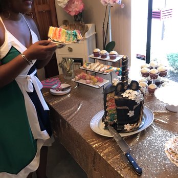 Babicakes - a woman standing in front of a table with a cake and cupcakes - Desserts Near Me Babicakes - a woman standing in front of a table with a cake and cupcakes - Desserts Near Me