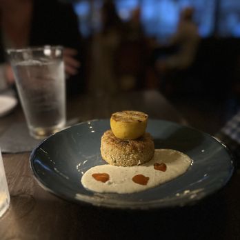 River & Post - Crab cake - A dish featuring a grilled fruit on top of a breaded or croquette-like base, accompanied by a white sauce with small drops of red sauce, served on a dark plate. - Lounges Near Me - Jacksonville, Florida