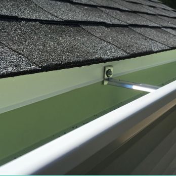 Ray Roofing - The image depicts a section of a roof with black asphalt shingles installed above a green gutter system. The gutter is supported by metal brackets attached to the fascia board. The shingles are textured and layered, while the gutter appears clean and well-maintained. The background shows a hint of greenery and a clear sky. - Roofing Near Me - Hackensack, New Jersey
