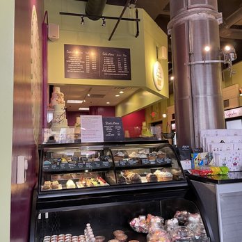 Classy Girl Cupcakes - a display of cupcakes - A bakery interior features a glass display case filled with cupcakes, cakes, and bottled beverages. Behind the case, a menu board lists various cupcake flavors and prices. The background shows part of the bakery's decor, including a cake on a stand and a colorful wall. The setting appears to be a retail bakery offering a variety of baked goods and drinks. - Bakeries Near Me - Milwaukee, Wisconsin