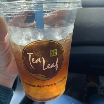 Tea Leaf Cafe - Strawberry kiwi iced tea with kiwi bobas - A person is holding a plastic cup of iced tea with a blue straw inside a car. The cup has a 'Tea Leaf Cafe' logo and the slogan 'Everyone deserves a little TLC.' The background shows the car's interior and a view of the sky and trees outside. - Coffee & Tea Near Me - Amherst, New York