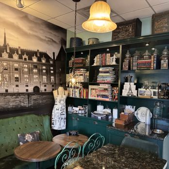 Cafe de Nook - The image shows the interior of a cafe with a large black-and-white mural of a cityscape on the wall, featuring historic buildings and spires. There are vintage chandeliers hanging from the ceiling, and a dark green bookshelf filled with books, decorative objects, and framed photos. A mannequin with a dress form stands near a green upholstered booth with a small round wooden table. The space has a cozy, vintage-inspired decor with a mix of modern and antique elements. - Coffee & Tea Near Me - Columbus, Ohio