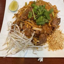 Pad Thai - A serving of Pad Thai on a white square plate, garnished with bean sprouts, crushed peanuts, cilantro, and a lime wedge, served on a wooden table.