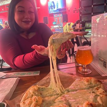 Voodoo Brewing - A woman with dark hair and makeup sits at a table in a brewery, holding a slice of cheese pizza with melted cheese stretching. - Brewpubs Near Me - Valrico, Florida