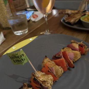 Nando's Peri-Peri - Grilled vegetable skewers with tomatoes, onions, and herbs are presented on a gray plate, accompanied by drinks and other dishes in the background. - South African Near Me - Wheaton, Maryland