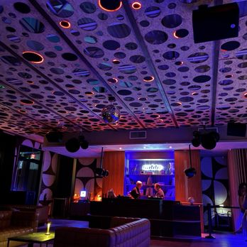 Sable - The image shows a nightclub interior with a ceiling made of a perforated material with numerous circular cutouts, illuminated by colorful lights. The space includes a DJ booth at the back with two individuals working behind it, and a neon sign above the booth. The room has lounge seating with leather couches and a small table, and the overall lighting creates a vibrant, modern atmosphere. - Bagels Near Me - Miami, Florida