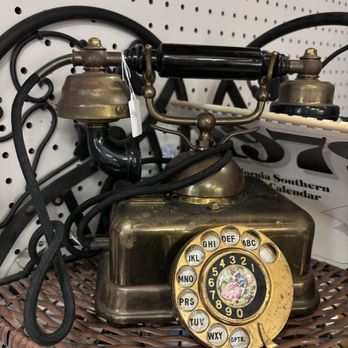Antiques On Pierce - The image shows an antique rotary dial telephone with a brass-colored body and a black handset. The rotary dial features a decorative center with a floral design and numbers from 1 to 9 and 0. The phone is placed on a woven wicker surface, with a pegboard wall and some black cords visible in the background. - Antiques Near Me - Milwaukee, Wisconsin