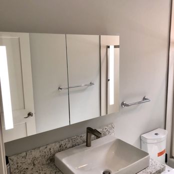 Poseidon Remodeling - The image shows a bathroom vanity with a rectangular white vessel sink installed on a marble countertop. Above the sink is a large mirror with vertical lighting fixtures on both sides. The wall behind the mirror is painted in a neutral color. To the right of the sink, a toilet is partially visible. The setup appears modern and minimalistic, with functional fixtures and clean lines. - General Contractors Near Me - Oceanside, California