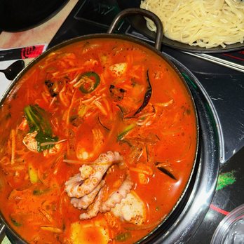 Goshen - spicy seafood soup hotpot (jjamppong) - A hotpot filled with a spicy red broth containing seafood such as octopus, along with vegetables like zucchini and green onions. The hotpot is placed on a table with a side of plain cooked noodles in a black dish. The setting appears to be a restaurant table with utensils and a glass of water nearby. - Karaoke Near Me - Glenview, Illinois