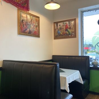 Curry Leaf - Booths (there are 4 in the restaurant) - Interior of a restaurant featuring booth seating with black leather upholstery and a white tablecloth. Two framed artworks depicting traditional Indian or Pakistani scenes hang on the white wall. A ceiling light illuminates the space, and a window shows a view of the outside street with cars and signage. - Indian Near Me - Foxborough, Massachusetts