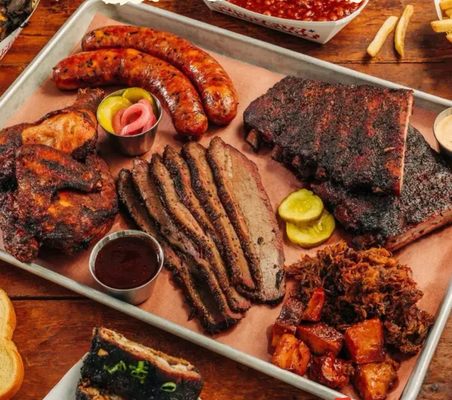 A large platter featuring a variety of smoked meats including brisket, ribs, sausage, and chicken, along with pickles and sauces.
