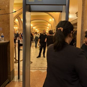 Auditorium Theatre - A metal detector gate is set up at the entrance of a historic theater, with people walking through or around it. The theater interior has arched ceilings, decorative lighting fixtures, and intricate architectural details. The scene includes individuals in formal or semi-formal attire, indicating an event or performance. The environment appears busy, with multiple attendees visible in the background. - Performing Arts Near Me - Chicago, Illinois