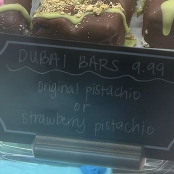 Baskin-Robbins - Dubi ice cream bar - Chocolate-covered ice cream bars are displayed on a stick, topped with green pistachio drizzle and sprinkled with crushed pistachio nuts. The sign indicates they are called Dubai Bars, priced at $9.99, with options for original pistachio or strawberry pistachio flavors. The image shows the bars arranged on a tray, with a handwritten sign in front of them. - Desserts Near Me - Modesto, California