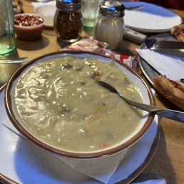 Green Chile Chicken Soup - A bowl of green chile chicken soup with a creamy texture and visible ingredients is on a restaurant table. The table also has condiments, glasses of water, pizza slices, and utensils, indicating a casual dining setting. The soup appears hearty and flavorful, typical of New Mexican cuisine.