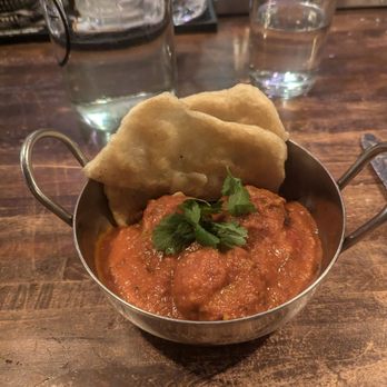 Thistle & Leek - A metal dish with Indian curry topped with cilantro and accompanied by naan bread is on a wooden table, with glasses of water and a glass pitcher in the background. - Gastropubs Near Me - Newton, Massachusetts