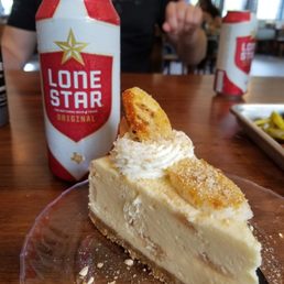 Banana Pudding Cheesecake - A slice of cheesecake topped with banana pieces and whipped cream is on a clear plate, with Lone Star beer cans and a person in the background.