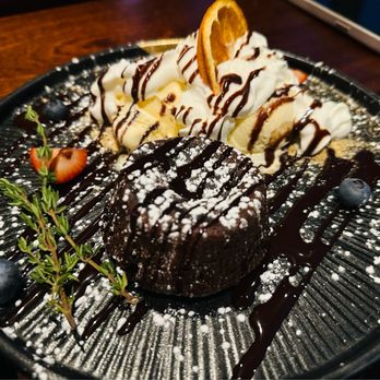 Tavern 157 - A dessert featuring a chocolate lava cake dusted with powdered sugar, served with whipped cream, banana slices, and garnished with blueberries and a sprig of herbs. - Lounges Near Me - Flushing, New York