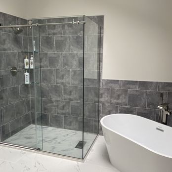 Three Rivers Builders - brand new master bathoroom - This bathroom showcases a glass shower enclosure with gray tiled walls and a marble floor, equipped with a showerhead and built-in shelves holding toiletries. Adjacent to the shower is a white freestanding bathtub with a modern faucet, set against a wall with matching gray tiles. The space has a minimalist design with neutral tones and clean lines. - Damage Restoration Near Me - Severna Park, Maryland