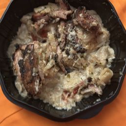 Supposedly The Jerk Chicken Alfredo - A black takeout container holds a creamy, cheesy dish with grilled chicken pieces on top, served on an orange surface.