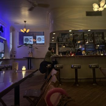 King Tai - Ambience - A bar interior featuring a curved bar counter with bar stools, ambient lighting, and a shelf with bottles and decorations behind the bar. - Cocktail Bars Near Me - Brooklyn, New York