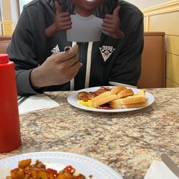 A person with a cartoon avatar mask featuring dreadlocks, a smiling face, and thumbs-up gestures is sitting at a restaurant table. The table has plates of breakfast food, including toast, scrambled eggs, and potatoes with ketchup. The background shows a typical restaurant interior with beige walls, a clock, and a window with blinds.