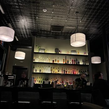 Kettlestrings Grove - Ambiance - The image shows an indoor bar with a dark, patterned ceiling and three hanging pendant lights. Behind the bar, illuminated shelves display a variety of liquor bottles in different colors and sizes. There are three bartenders or staff members visible, and a few patrons seated at the bar. The overall ambiance appears dimly lit and modern. - Cocktail Bars Near Me - Oak Park, Illinois