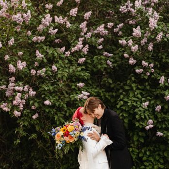 Riley Glenn Photography - Two non binary marriers embracing in front of flower bush - Session Photography Near Me Riley Glenn Photography - Two non binary marriers embracing in front of flower bush - Session Photography Near Me