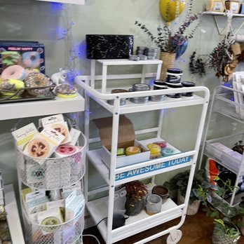 Raw Edge - A white metal rolling cart with three shelves contains small containers, potted plants, and decorative objects. The store has shelves on the wall displaying various items, including vases, framed pictures, and decorative pieces. The background features a light green wall with wall-mounted shelves and hanging decor, including dried branches and vases. The floor is wooden, and the overall setting appears to be a boutique or gift shop with diverse home decor and gift items. - Flowers & Gifts Near Me - Chicago, Illinois