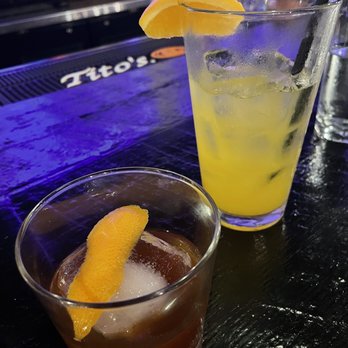 Free State - CC Old Fashioned and Orange Crush both absolutely amazing cocktails! - The image shows two cocktails on a dark bar counter, each garnished with a lemon wedge. The taller glass contains a yellow-colored drink with ice and a straw, while the shorter glass holds a darker beverage with ice. The background features bar equipment and bottles, indicating a bar setting. - Wine Bars Near Me - Washington, DC, District of Columbia