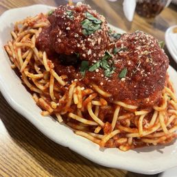 Meatballs - A serving of spaghetti pasta topped with two large meatballs covered in tomato sauce, garnished with chopped herbs and grated cheese, presented in a white bowl on a wooden table. The background includes a white cup, a jar of spices, and some napkins, indicating a restaurant environment.