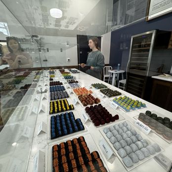 Splendid Oaks Chocolates - Chocolates - Assorted chocolates are arranged on a white marble counter inside a chocolate shop, with a glass display and two customers observing. - Chocolatiers & Shops Near Me - Waco, Texas