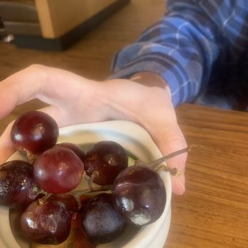 Denny's - MOLD ON MY GRAPES! - A person wearing a blue plaid shirt is holding a small white bowl containing red and black grapes. The background shows a wooden table and part of the restaurant interior with warm lighting and wooden flooring. - Diners Near Me - Bastrop, Texas