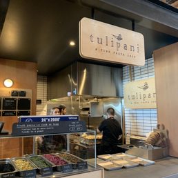 Inside Tulipani, a tiny pasta shop, there is a counter with fresh pasta ingredients and a menu board, with staff preparing food.