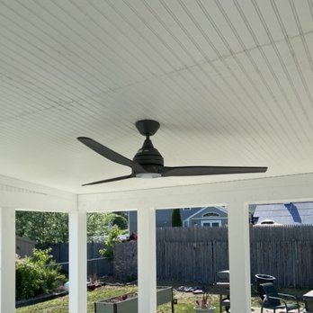 Drohan Development - The image shows a black ceiling fan installed on a white, paneled ceiling of a covered outdoor patio. The patio has white support columns and offers a view of a backyard with a wooden fence, garden plants, and outdoor furniture including chairs and a table. The scene is illuminated by natural daylight, with neighboring houses visible in the background. - General Contractors Near Me