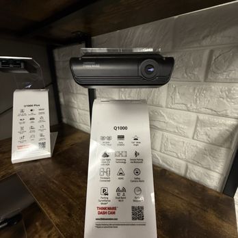 Ezdashcam - A black Thinkware dash cam is attached to a wooden shelf, positioned against a white brick wall. A product information tag hangs from the device, listing features such as 2K resolution, 156° wide angle, HDR, Bluetooth pairing, and parking surveillance. The setting appears to be a retail display showcasing dash cam models and accessories. - Auto Security Near Me - Tempe, Arizona