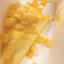 Mango Mille Crepe Cake - A slice of mango crepe cake with multiple thin layers is shown inside a white box. The cake is topped with diced mango pieces and a dusting of powdered sugar. Some mango chunks and crumbs are scattered around the cake inside the box, which appears to be a dessert item from Mango Mango Dessert in Plainview, NY.