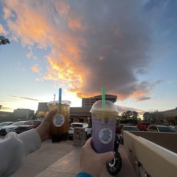 Rice Box & Boba  - Two beverages, one dark and one purple, are held by hands in front of a parking lot and a sunset sky at Rice Box & Boba in Albuquerque, NM. - Laotian Near Me - Albuquerque, New Mexico