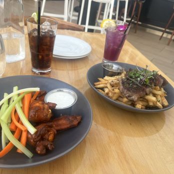 Offshore Rooftop - Steak frites & chicken wings. - A table with two plates of food: chicken wings with vegetable sticks and steak fries with grilled meat, along with drinks including iced tea and a purple cocktail. - New American Near Me - Chicago, Illinois