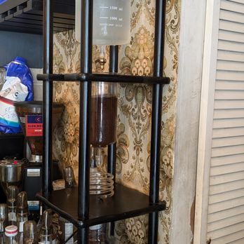 OK Cafe - A coffee siphon brewing device with a glass chamber containing dark coffee, supported by a black metal frame on a shelf. The device includes a water chamber at the top and a lower chamber with brewed coffee. The background features patterned wallpaper and various coffee supplies, indicating a cafe environment. - Coffee & Tea Near Me - Astoria, New York