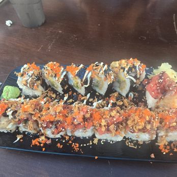 Dragon Sushi - Rolls! - A black rectangular plate features an assortment of sushi rolls, including some with fish roe, drizzled sauces, and crunchy toppings. The rolls are arranged in two rows, with some garnished with orange fish roe, white sauce, and sesame seeds. A small portion of wasabi is placed on the side of the plate. The plate is set on a dark wooden table, with a cup, a napkin, and some other items visible nearby. - Sushi Bars Near Me - Albuquerque, New Mexico